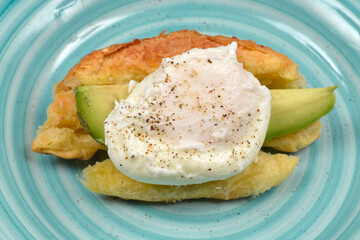 A croissant is sliced and filled with avocado slices and a poached egg, served on a textured blue plate