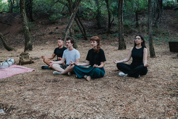 A small group sits cross legged on the forest ground, eyes closed, practicing mindfulness and...