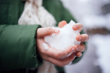 Person holding a snowball in hands, wearing a green winter jacket and a light scarf, surrounded by a snowy landscape, capturing the essence of winter fun and playfulness