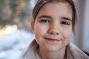 Young girl with brown hair and blue eyes, smiling warmly, wearing a cozy sweater, with a softly blurred winter background, capturing a moment of joy and innocence