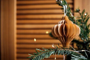 Decorative paper ornament hanging from a pine branch, with warm wooden background and soft glowing lights, creating a cozy festive atmosphere for holiday celebrations