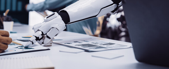 A robotic arm assists a professional in a modern office, focusing on data analysis and charts. This image highlights the blend of technology and innovation in the workspace.Noogenesis