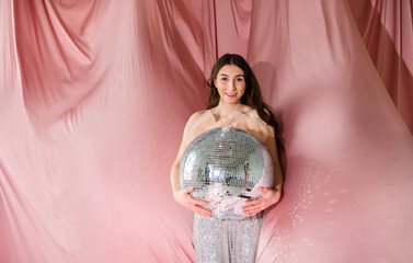 Young woman with long dark hair, wearing a sparkling outfit, joyfully holding a large disco ball against a soft pink backdrop, embodying celebration and festive spirit