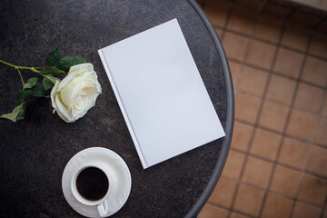 White rose and coffee cup placed on a dark table beside an empty notebook, creating a serene atmosphere for reflection and creativity in a cozy setting