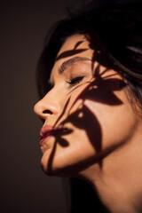Profile of a woman with dark hair, illuminated by soft light, showcasing delicate shadows cast by plant leaves, creating a serene and artistic atmosphere in a minimalist setting