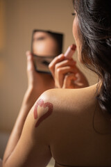 Woman applying lipstick while looking in a small mirror, showcasing a heart-shaped design on her shoulder, reflecting beauty and self-expression in a serene setting
