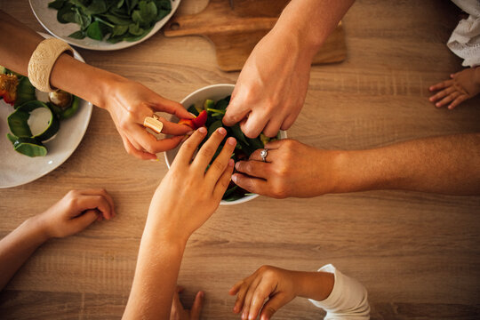 Diverse hands of adults and children reaching for fresh salad ingredients in a bowl on a wooden table, showcasing family bonding and healthy eating habits - Powered by Adobe