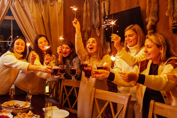 Young women in knitted sweaters laugh and raise glasses of mulled wine. Friends have fun and drink wine with spices. New Year Eve party with sparklers.