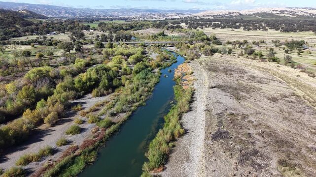 Drone flies forward above Cache Creek toward a small bridge, showing dense vegetation, dry banks and distant hills under a clear sky in Capay, California.
