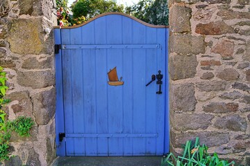 Charming blue wooden gate with sailboat detail stands between weathered stone walls, inspiring dreams of summer coastal getaways and nautical adventures