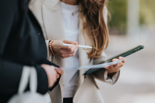Two young professionals stand outdoors, examining printed documents while one holds a tablet and stylus.