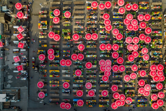 Aerial view of a vibrant marketplace with rows of stalls sheltered by red umbrellas casting shadows on the ground, Zagreb, Croatia.