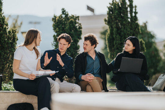 A diverse group of young professionals sit outdoors, discussing ideas while sharing a laptop and papers in a sunny, relaxed work session.