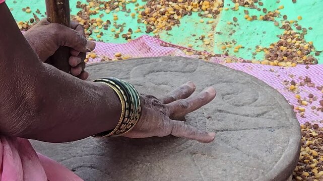 A woman uses a traditional hand-cranked stone mill ("chakki") to grind chickpeas. Fingers clear stuck grains from the central hole, maintaining the manual grinding process to make chickpea pulses.