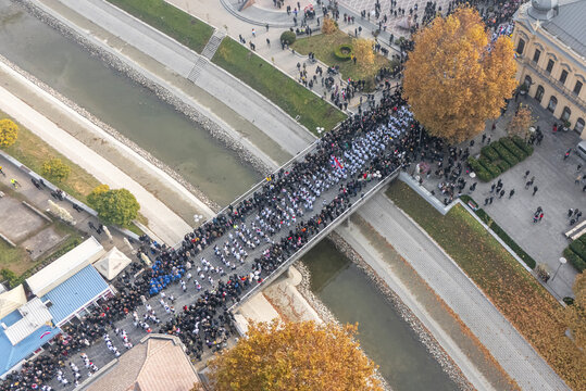 Vukovar, Croatia - 18 November 2025: Aerial view of a somber procession crossing a bridge over the Vuka River, flanked by historical buildings and solemn onlookers.