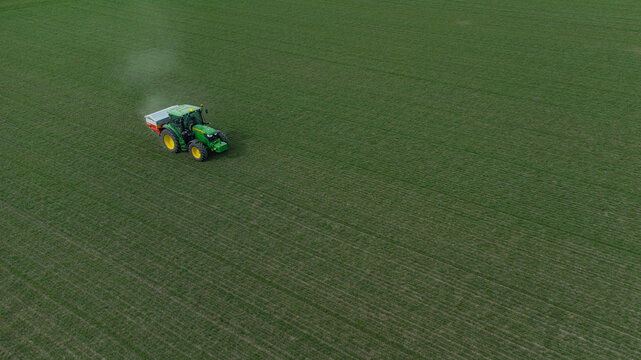 Aerial view of a vibrant green tractor spreading fertilizer over the lush fields, creating a stark contrast against the soil, Vladislavci, Osijek-Baranja County, Croatia.