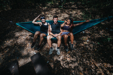 Three friends rest in a blue hammock hung between trees on a sun-dappled forest floor. Casual athletic wear, relaxed poses, and a moment of companionship during a nature retreat.
