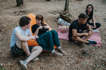 A diverse group of friends relaxes during a forest camping trip, chatting and enjoying a moment on a checkered blanket with a picnic basket and backpacks nearby.