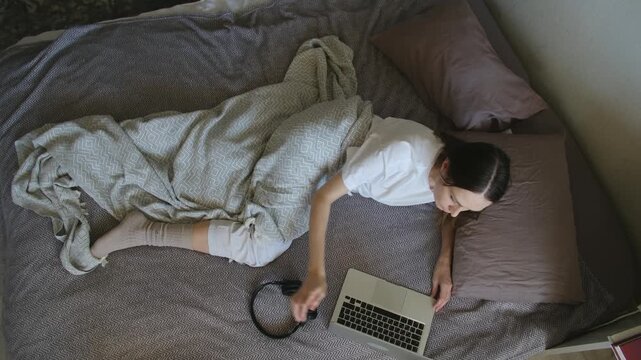 Tired young woman working on her computer with headphones in bed, eventually taking them off, closing laptop, and falling asleep from exhaustion, viewed from high angle in her bedroom at day time.