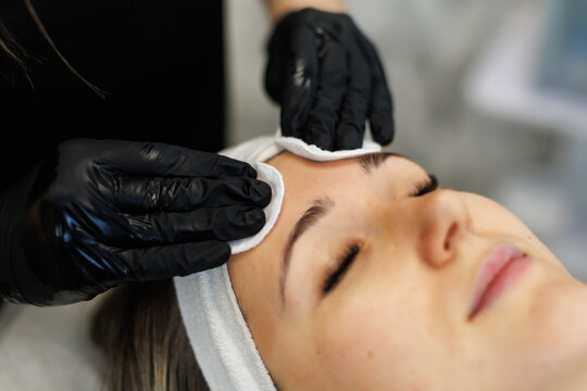 A close-up of a facial treatment showing a beautician in black gloves gently cleansing a client's forehead with cotton pads during a spa session