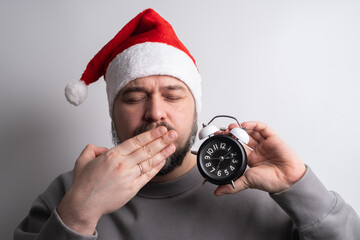 Exhausted man in santa hat yawning holding alarm clock