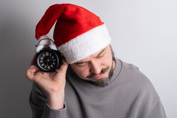 Exhausted man in santa hat yawning holding alarm clock
