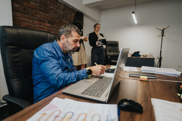 A businessman is working at their desk while colleagues engage in discussion. The office setting reflects a collaborative work environment, showcasing productivity and professional engagement.