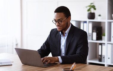 Fototapeta premium Cheerful young entrepreneur in formal suit working with laptop, wearing glasses, office interior, copy space. Successful millennial african american businessman working on business project