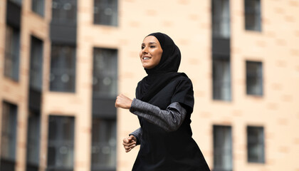 Glad confident young muslim woman in black hijab and sportswear runs outdoors, on building background, free space. Vitality, urban workout season, jogging, sports in city, health care and weight loss