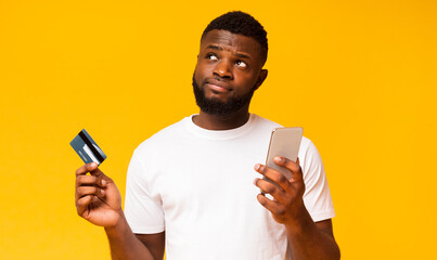Do I longer need a credit card. Hesitant african american man holding smartphone and card, looking upwards