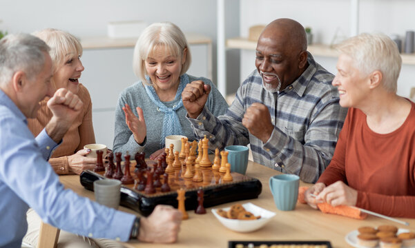 Emotional multiethnic group of senior people playing chess, happy elderly men and women in casual outfits sitting around table with table games, chatting, having fun at sanatorium