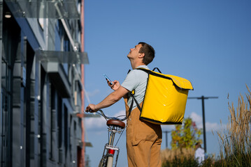 Bike courier wearing a yellow delivery bag and brown overalls, holding a smartphone and looking up; navigating city streets for food delivery in an urban setting