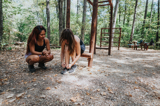 Two women crouch by a wooden exercise frame in a forest setting, tying sneakers and sharing a casual moment during an outdoor activity. - Powered by Adobe
