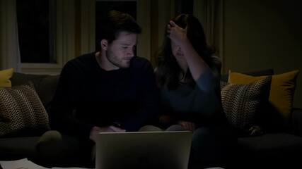 Young couple sitting on a couch at home late at night, reviewing bills and financial documents, looking worried and focused on a laptop screen while calculating expenses - Powered by Adobe