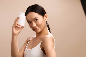 Asian woman in a simple studio pose holds a white bottle, presenting a clean lifestyle product. She smiles softly, exuding calm confidence and modern elegance, creating a wellness and beauty oriented
