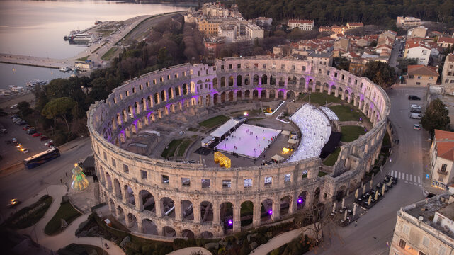 Aerial view of the ancient Roman amphitheater, now an ice rink, glows warmly against the cool twilight, contrasting with the calm sea in the background, Pula, Istria County, Croatia.