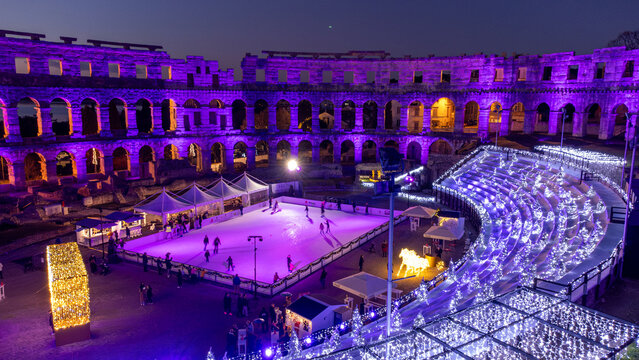 Pula, Croatia - 17 November 2025: Aerial view of the ancient Roman amphitheater illuminated in vibrant purple hues, hosting a bustling ice rink amidst a sea of twinkling lights.