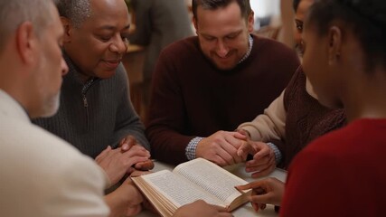 Intergenerational and multi ethnic community group sitting together at a table, holding hands and intently reading from an open bible during a quiet spiritual gathering