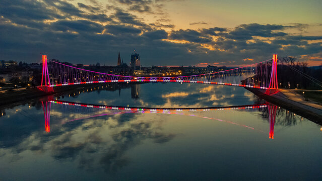 Aerial view of the illuminated pedestrian bridge reflecting vividly over the water, with the cathedral and city lights softly glowing in the background, Osijek, Osijek-Baranja County, Croatia. - Powered by Adobe