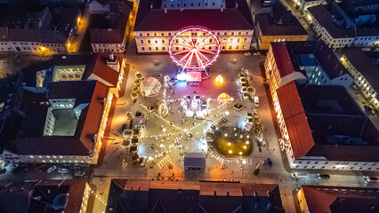 Aerial view of a brightly illuminated square featuring a Ferris wheel and festive lights nestled between buildings, creating a warm, inviting glow, Osijek, Osijek-Baranja County, Croatia.