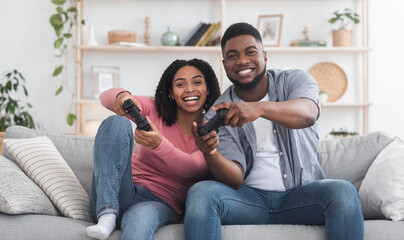 Obraz premium Cheerful African American couple competing with each other in video games, holding joysticks and laughing, staying at home