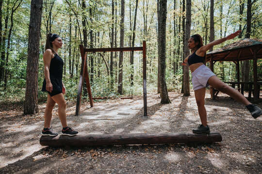 Two women exercise together in a wooded park, balancing on a wooden beam as part of an outdoor obstacle course. Bright sunlight, nature backdrop, teamwork and motivation in a fitness moment. - Powered by Adobe