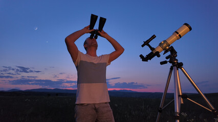 Fototapeta premium Amateur astronomer using telescope and binoculars for watching stars, Moon, planets and other celestial objects from a countryside field in nature.