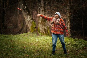 A boy in a red plaid jacket and scarf throws a red discus in a grassy park with trees. Concentrated outdoor activity in cool weather.