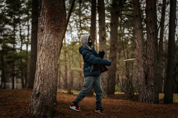Fototapeta premium A boy in a gray hoodie and blue puffer jacket throws a disc on a grassy field with a forest backdrop. Dynamic action shot during cool weather.