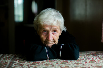 An elderly woman rests her head on folded arms at a patterned table, her gentle expression softened...