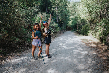 Two women with backpacks and trekking poles share a cheerful moment on a forest gravel trail, enjoying a sunny day and outdoor adventure together.