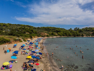 Korcula, Croatia - 14 November 2025: Aerial view of a sun-kissed beach teeming with vibrant umbrellas and bathers, nestled between the tranquil, clear waters and the lush green hills under a blue sky.