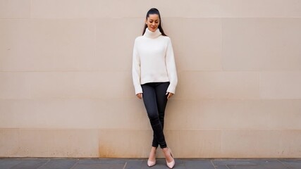 Woman in white turtleneck and black pants leans against a light stone wall
