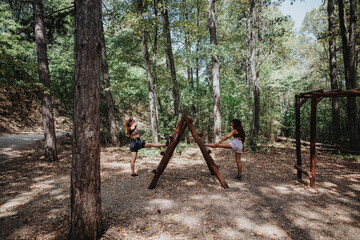 Two friends workout outdoors in a forest park, balancing on a wooden obstacle frame as they kick and stretch. The sunny, leafy setting conveys fitness, teamwork, and outdoor adventure.